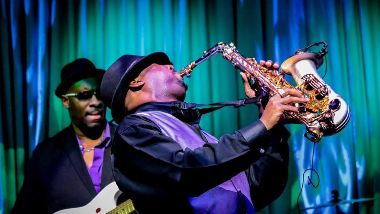 Seorang musisi jazz memainkan saksofon di atas panggung dengan suasana penuh cahaya hangat