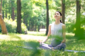 Seseorang duduk bersila dengan mata tertutup di tengah alam, menciptakan suasana tenang dan damai sebagai bentuk meditasi dan relaksasi untuk menenangkan pikiran.