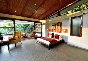 Interior kamar villa dengan sentuhan tradisional Bali, dilengkapi tempat tidur kayu, dekorasi etnik, dan balkon dengan pemandangan alam
