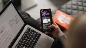 Seseorang menggunakan laptop sambil membaca berita di ponsel, mencerminkan kebiasaan konsumsi informasi yang berlebihan, yang dapat memengaruhi pola pikir dan kesehatan mental