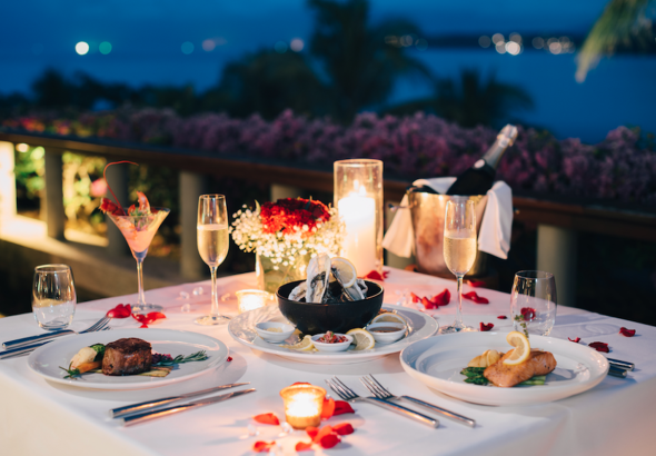 Tempat makan malam romantis dengan steak, seafood, dan sampanye, di teras yang menghadap laut pada malam hari.